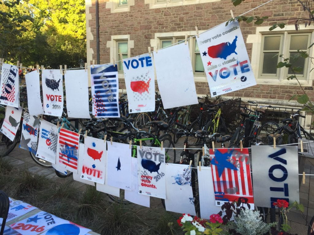 Spray-painted posters across multiple clotheslines in red, blue, silver, and black with stars, stripes, with patriotic, democratic language and iconography urging voting.