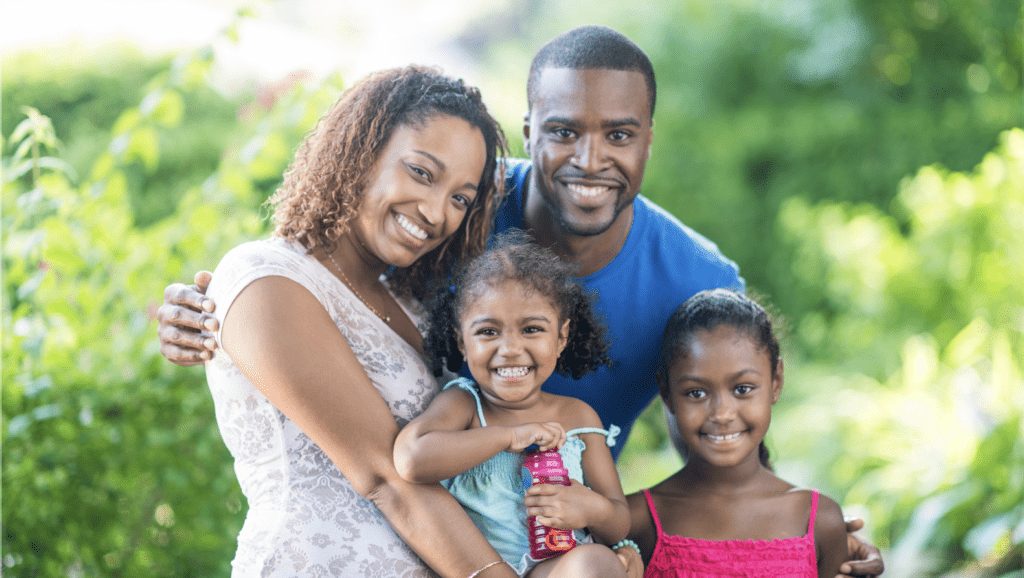 African American family of four with two young children, smiling at camera with greenery in background.