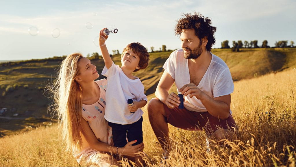 Male and female couple playing with young child playing with soap bubbles in nature.