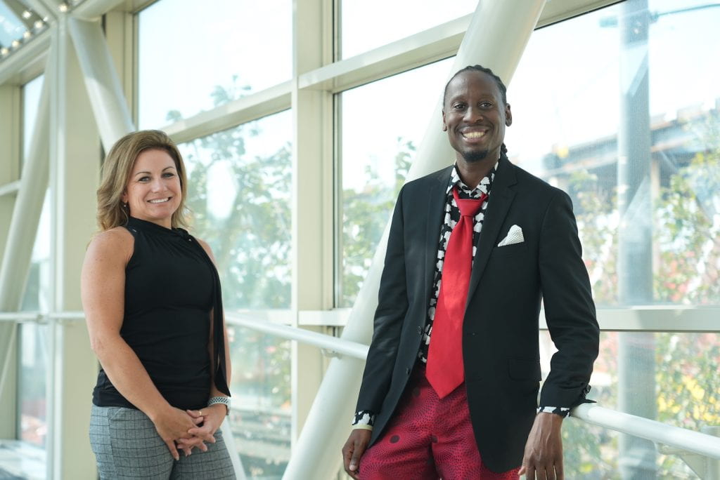 Two people in an indoor walkway with large windows, smiling at the camera. On left a white woman in black top. On right a black man in dark sport coat.