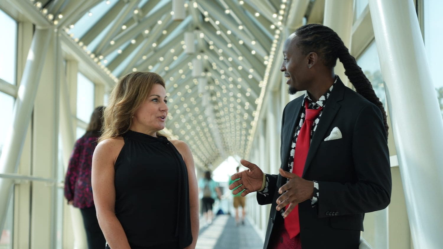 Two people in an indoor walkway with large windows, having a nice, animated discussion. On left a white woman in black top. On right a black man in dark sport coat.